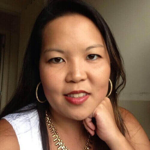 Mulher asiática de cabelo escuro e liso, usando brincos de argola e colar dourado, com a mão apoiada no queixo e vestindo blusa branca.